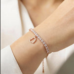 Rose gold tennis bracelet with square crystal stones and dangling open heart charm on a delicate chain.